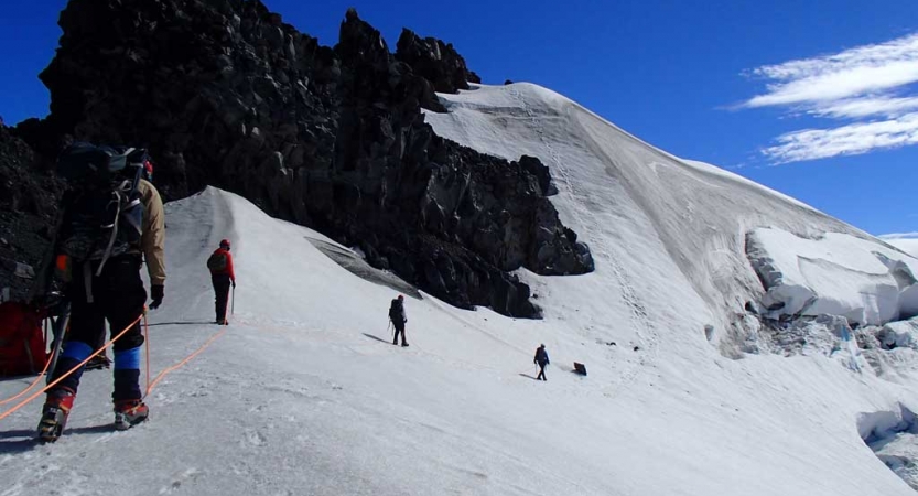 A group of people wearing safety gear are attached together by a rope, as they ascend a snowy, mountainous incline.